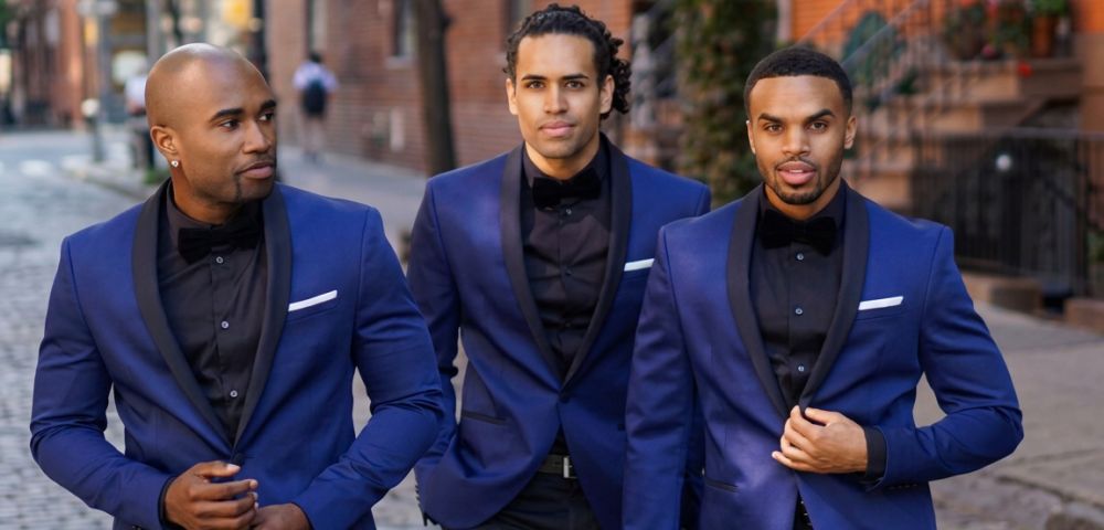 Three men in matching blue tuxedos with black lapels and bow ties stand on a cobblestone street.