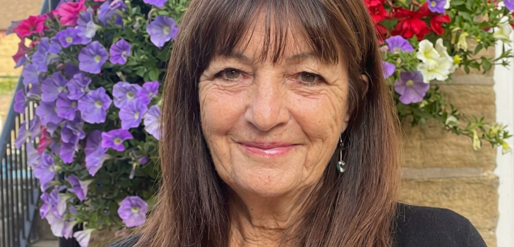 Close-up portrait of an older woman with long brown hair and bangs, smiling, with a hanging basket of purple and pink petunias behind her.