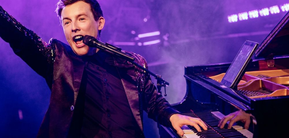 A male singer with short brown hair wears a shimmering dark jacket and sings into a microphone while playing a grand piano on a stage lit by purple lights.