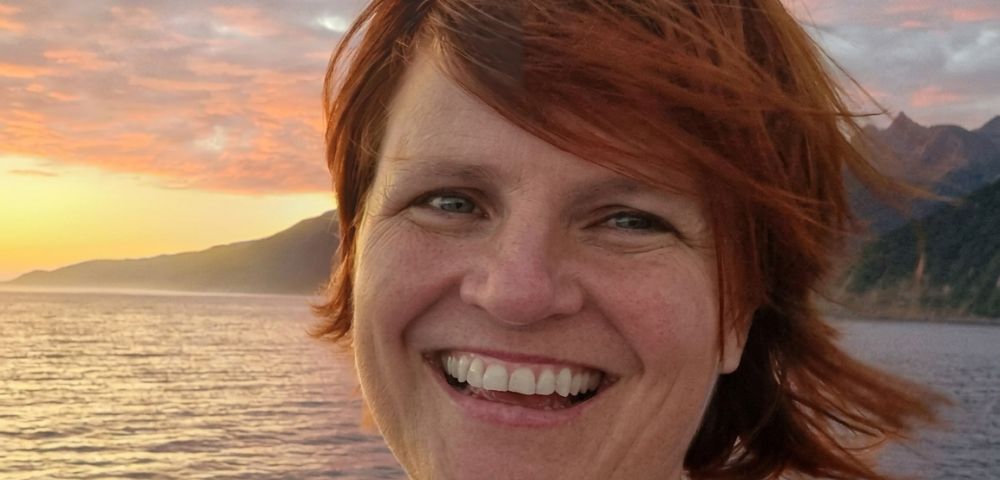 Close-up portrait of a woman with auburn hair smiling brightly, with a tranquil body of water, rolling hills, and a dramatic, colorful sunset sky in the background.