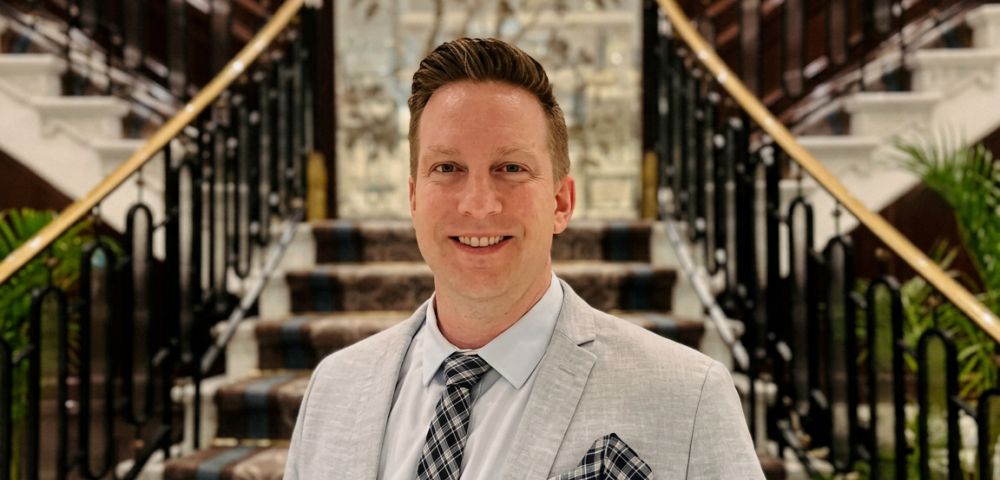 A man in a light grey suit jacket, collared shirt, and patterned tie smiles forward, standing on a carpeted staircase with dark ornate railings and a decorative wall behind him.