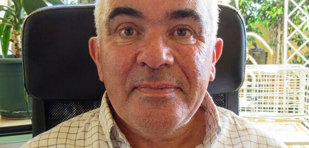 Close-up portrait of an older Caucasian man with short, graying hair and a checkered shirt, seated indoors with a plant and window in the background.