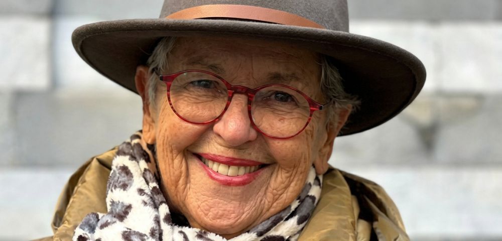 A smiling elderly person wearing a brown hat, red-rimmed glasses, a leopard-print scarf, and a tan jacket, with a blurred background of light-colored steps.