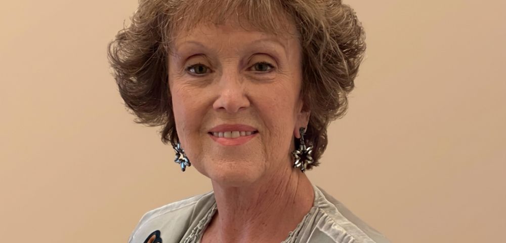 A medium shot of an older woman with short, curly brown hair and light skin, smiling at the camera. She is wearing a light-colored top with a detailed neckline and intricate, dark-colored earrings that resemble flowers or snowflakes. The background is a plain, light tan color.