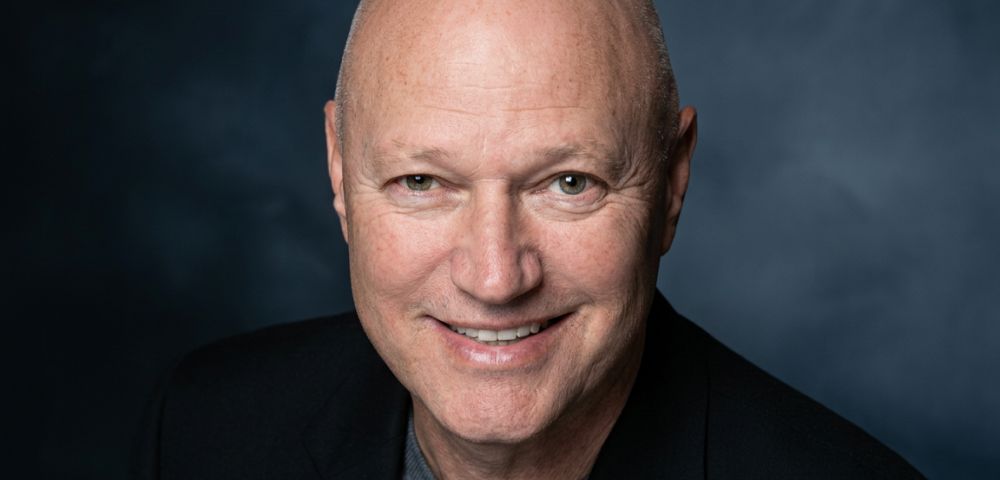 Close-up portrait of a bald, smiling man in a black suit jacket over a grey sweater against a dark, textured background.