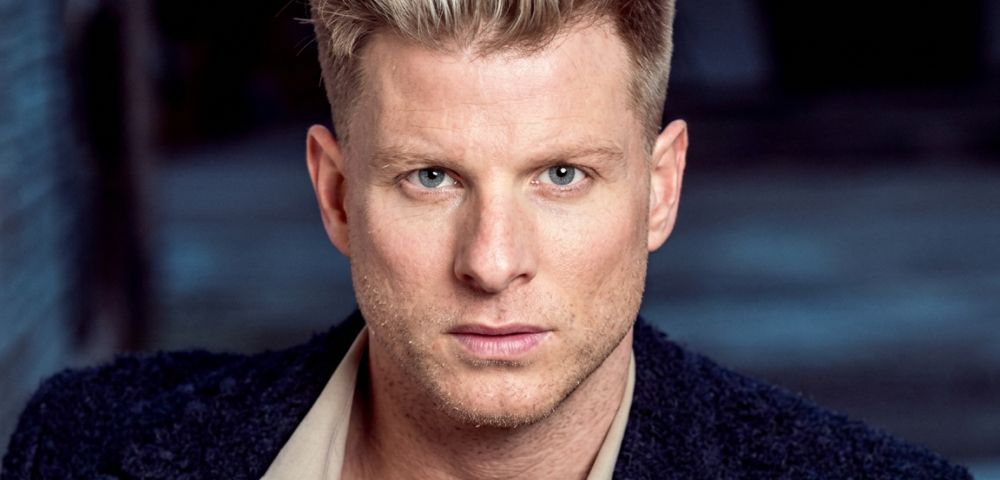 Close-up studio portrait of a man with blond spiky hair, light blue eyes, and stubble, wearing a beige collared shirt and a dark textured jacket.