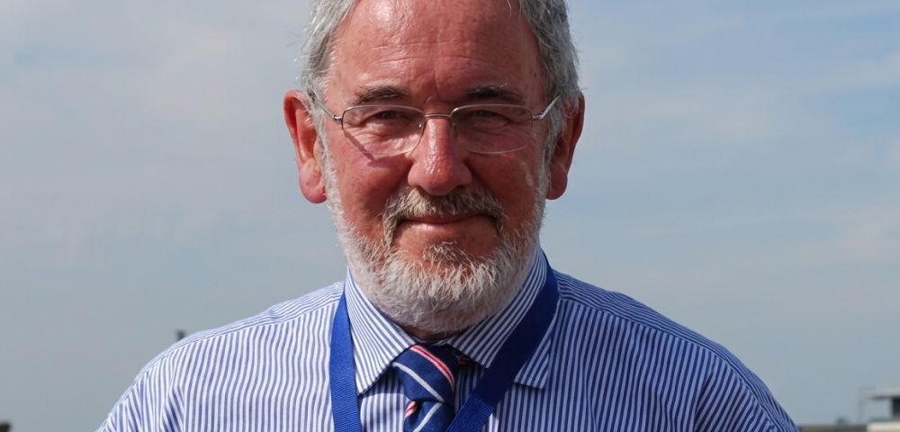 Close-up portrait of an older man with gray hair and a beard, wearing glasses, a striped blue shirt, and a tie. He is looking directly at the camera.