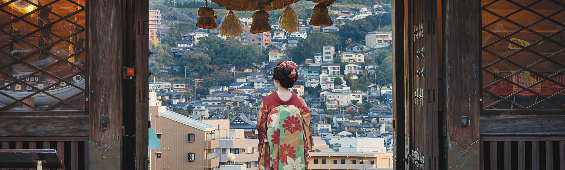 A person in a traditional Japanese kimono stands with their back to the viewer, looking out over a hillside city from within a large wooden gate adorned with a thick rope and straw ornaments.