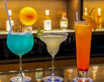 Three colorful cocktails with garnishes are displayed on a bar, with blurred bottles and a warm light in the background.