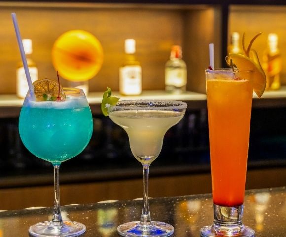 Three colorful cocktails with garnishes are displayed on a bar, with blurred bottles and a warm light in the background.