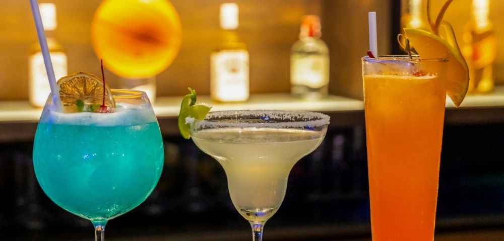 Three colorful cocktails with garnishes are displayed on a bar, with blurred bottles and a warm light in the background.
