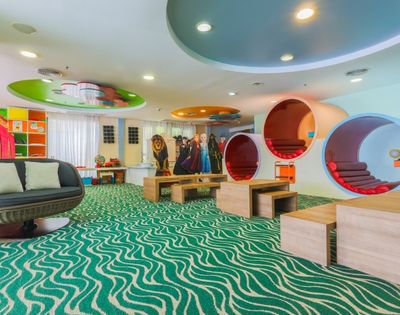 A brightly lit, colorful children's play area with a wavy green carpet, a circular outdoor-style sofa, and uniquely designed cylindrical pods for sitting and playing. Cardboard cutouts of characters and a lion stand behind wooden tables.
