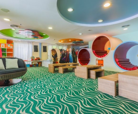 A brightly lit, colorful children's play area with a wavy green carpet, a circular outdoor-style sofa, and uniquely designed cylindrical pods for sitting and playing. Cardboard cutouts of characters and a lion stand behind wooden tables.