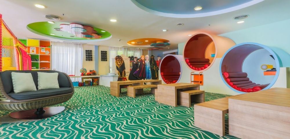 A brightly lit, colorful children's play area with a wavy green carpet, a circular outdoor-style sofa, and uniquely designed cylindrical pods for sitting and playing. Cardboard cutouts of characters and a lion stand behind wooden tables.