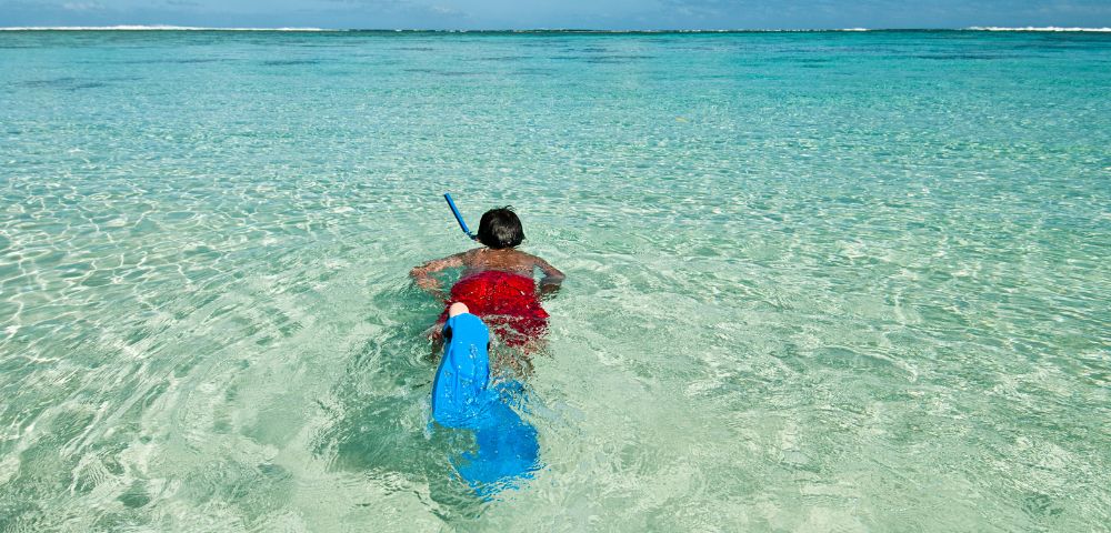A person wearing red swim trunks, a snorkel mask, and blue fins swims in crystal-clear turquoise ocean water under a bright blue sky with fluffy white clouds.