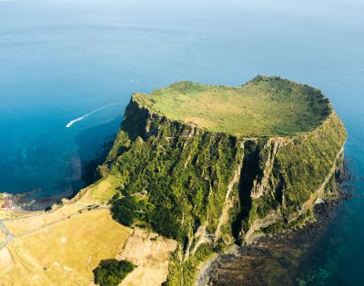 Aerial view of a volcanic crater island with steep, lush green cliffs rising from the deep blue sea, and a single white boat leaving a wake in the water.