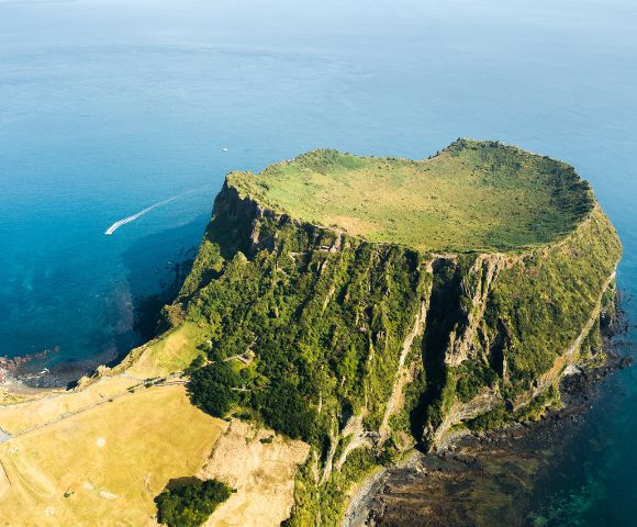 Aerial view of a volcanic crater island with steep, lush green cliffs rising from the deep blue sea, and a single white boat leaving a wake in the water.