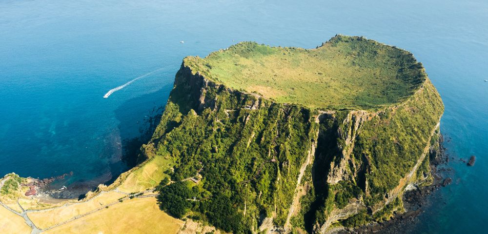 Aerial view of a volcanic crater island with steep, lush green cliffs rising from the deep blue sea, and a single white boat leaving a wake in the water.