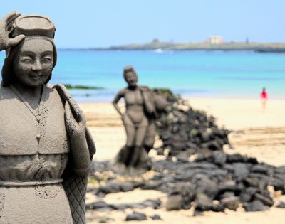 Stone statue of a woman with a headpiece and traditional clothing in the foreground, with a blurred statue of a woman carrying a burden and a distant figure on a sandy beach with the ocean and a faint island in the background.