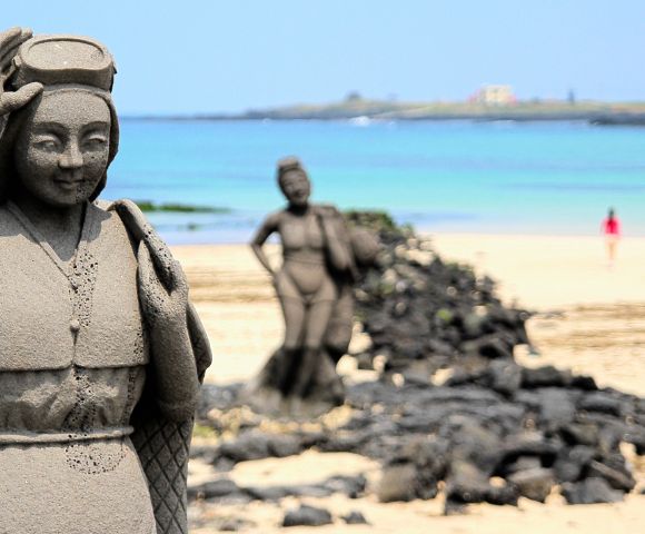 Stone statue of a woman with a headpiece and traditional clothing in the foreground, with a blurred statue of a woman carrying a burden and a distant figure on a sandy beach with the ocean and a faint island in the background.