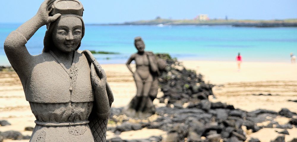 Stone statue of a woman with a headpiece and traditional clothing in the foreground, with a blurred statue of a woman carrying a burden and a distant figure on a sandy beach with the ocean and a faint island in the background.