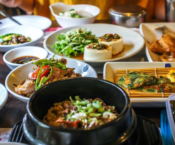 A variety of Korean dishes are arranged on a table, including a black stone pot filled with a savory meat and rice dish, a white bowl of sliced chicken with peppers, a plate of savory pancakes, and a dish of steamed tofu topped with seasonings.