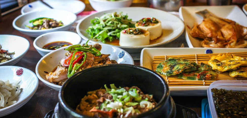 A variety of Korean dishes are arranged on a table, including a black stone pot filled with a savory meat and rice dish, a white bowl of sliced chicken with peppers, a plate of savory pancakes, and a dish of steamed tofu topped with seasonings.