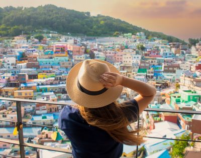 A person wearing a straw hat and a dark blue shirt looks out over a colorful hillside village with a mountain in the background at sunset.