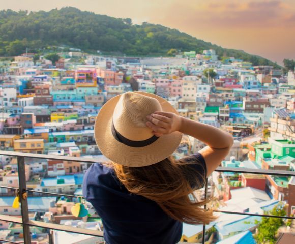 A person wearing a straw hat and a dark blue shirt looks out over a colorful hillside village with a mountain in the background at sunset.