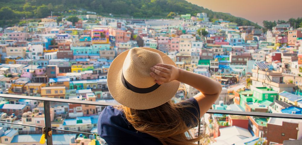 A person wearing a straw hat and a dark blue shirt looks out over a colorful hillside village with a mountain in the background at sunset.
