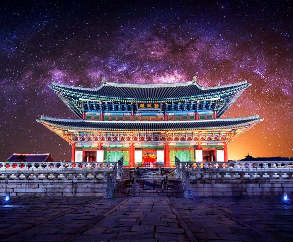 Traditional Korean palace building with ornate tiled roofs and colorful architectural details, illuminated at night against a vibrant purple and orange starry sky.
