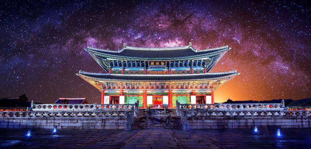 Traditional Korean palace building with ornate tiled roofs and colorful architectural details, illuminated at night against a vibrant purple and orange starry sky.