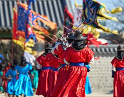 A procession of individuals in traditional Korean royal guard uniforms and wide-brimmed hats with feather accents, holding weapons, stand in front of colorful ceremonial flags at a historical site.