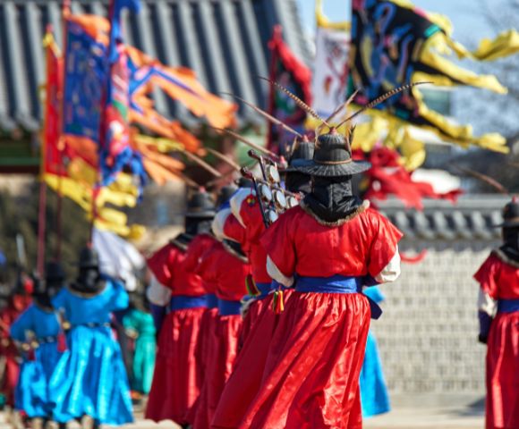 A procession of individuals in traditional Korean royal guard uniforms and wide-brimmed hats with feather accents, holding weapons, stand in front of colorful ceremonial flags at a historical site.