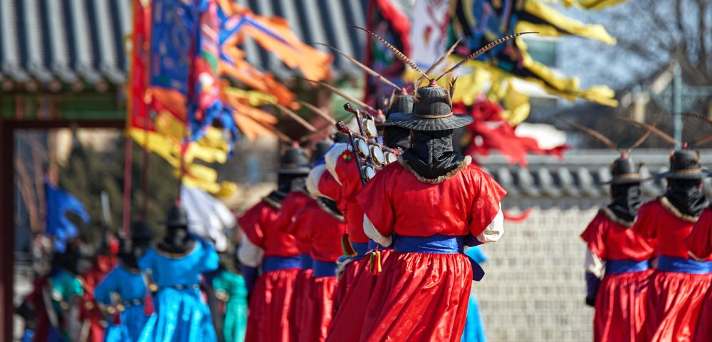 A procession of individuals in traditional Korean royal guard uniforms and wide-brimmed hats with feather accents, holding weapons, stand in front of colorful ceremonial flags at a historical site.