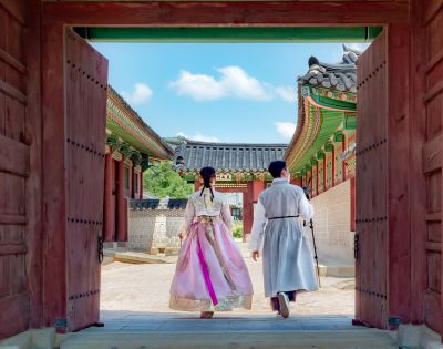 Two individuals in traditional Korean attire, a woman in a pink hanbok and a man in a light grey hanbok, walk through a large wooden gate into a courtyard of a traditional Korean palace with ornate roofs and colorful details under a bright blue sky.