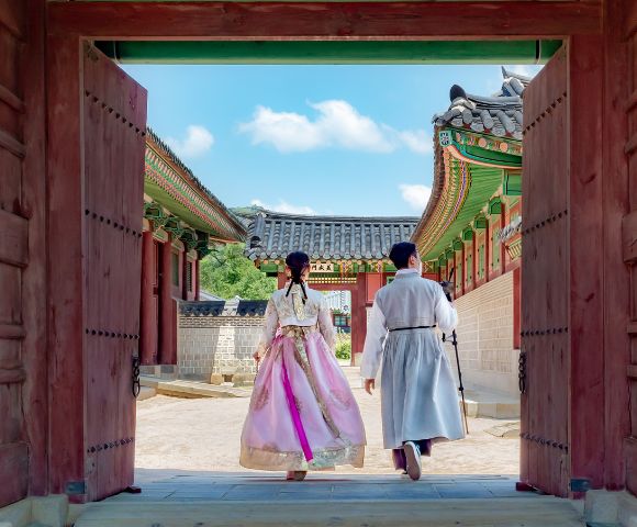 Two individuals in traditional Korean attire, a woman in a pink hanbok and a man in a light grey hanbok, walk through a large wooden gate into a courtyard of a traditional Korean palace with ornate roofs and colorful details under a bright blue sky.