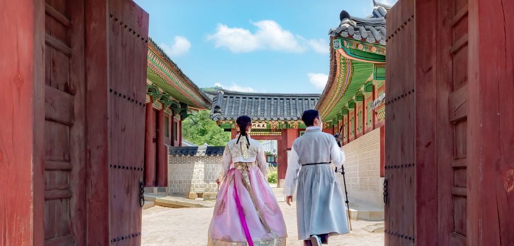 Two individuals in traditional Korean attire, a woman in a pink hanbok and a man in a light grey hanbok, walk through a large wooden gate into a courtyard of a traditional Korean palace with ornate roofs and colorful details under a bright blue sky.