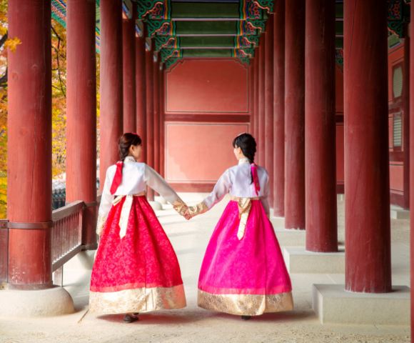 Two individuals wearing traditional Korean hanbok dresses, one in red and the other in bright pink, walk hand-in-hand down a covered walkway with large red pillars.