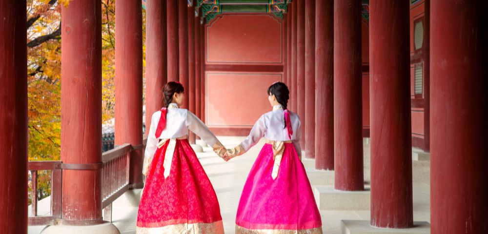 Two individuals wearing traditional Korean hanbok dresses, one in red and the other in bright pink, walk hand-in-hand down a covered walkway with large red pillars.