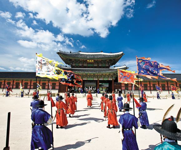 A ceremonial procession of individuals in traditional Korean royal guard uniforms and elaborate flags moves across a courtyard towards a grand palace gate under a bright blue sky with scattered clouds.