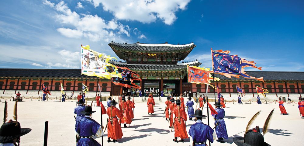 A ceremonial procession of individuals in traditional Korean royal guard uniforms and elaborate flags moves across a courtyard towards a grand palace gate under a bright blue sky with scattered clouds.