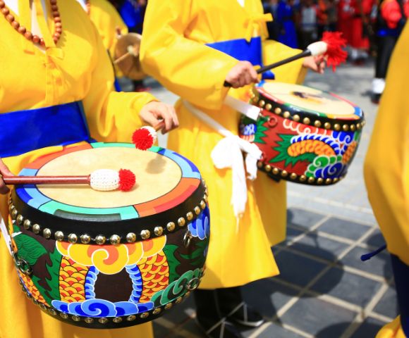 Two people in yellow robes with blue sashes play traditional drums decorated with colorful dragon motifs.