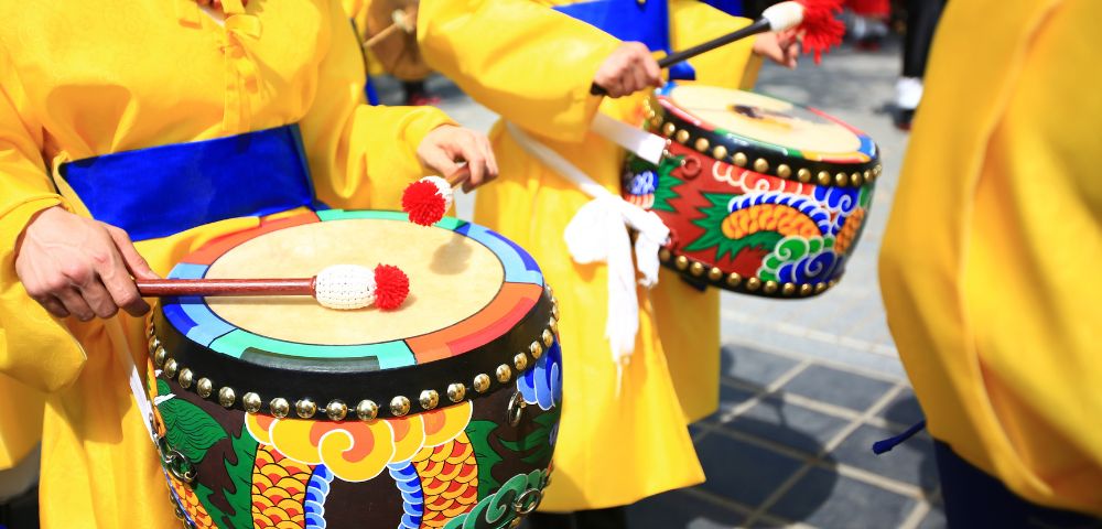 Two people in yellow robes with blue sashes play traditional drums decorated with colorful dragon motifs.