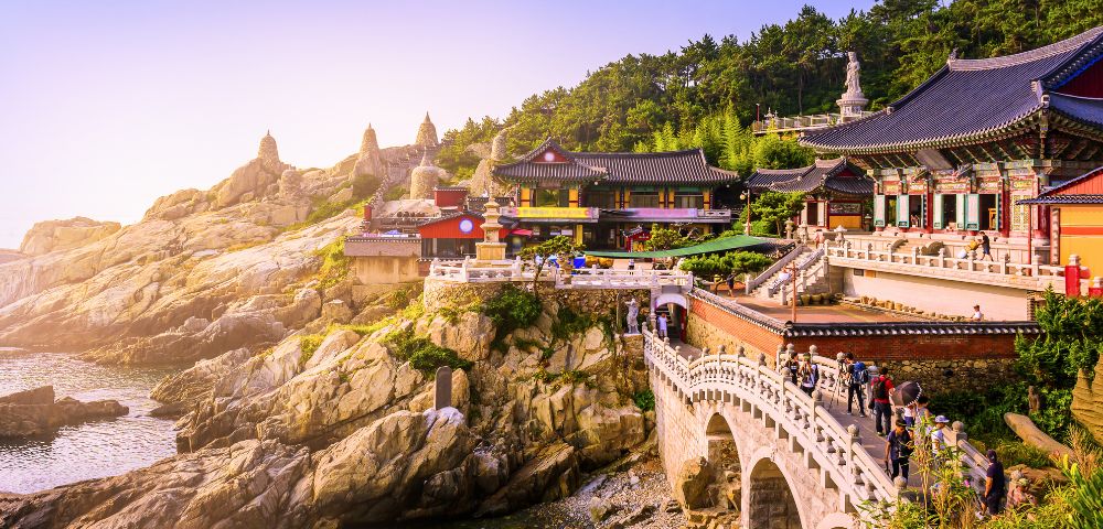 A scenic view of a colorful Buddhist temple complex nestled on a rocky cliff overlooking the ocean, connected by a bridge with people crossing.