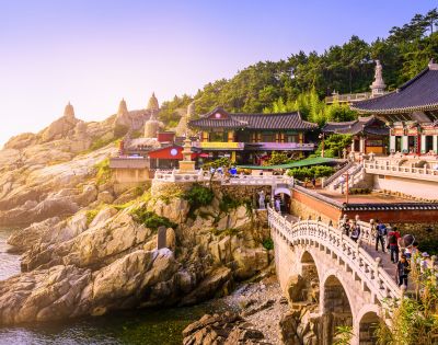 A scenic view of a colorful Buddhist temple complex nestled on a rocky cliff overlooking the ocean, connected by a bridge with people crossing.