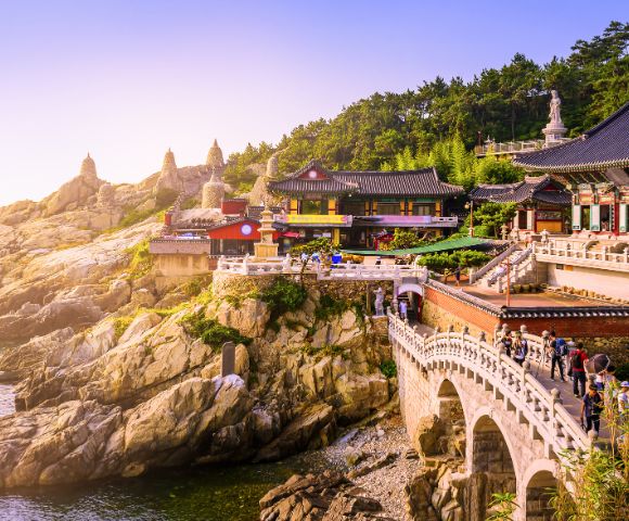 A scenic view of a colorful Buddhist temple complex nestled on a rocky cliff overlooking the ocean, connected by a bridge with people crossing.