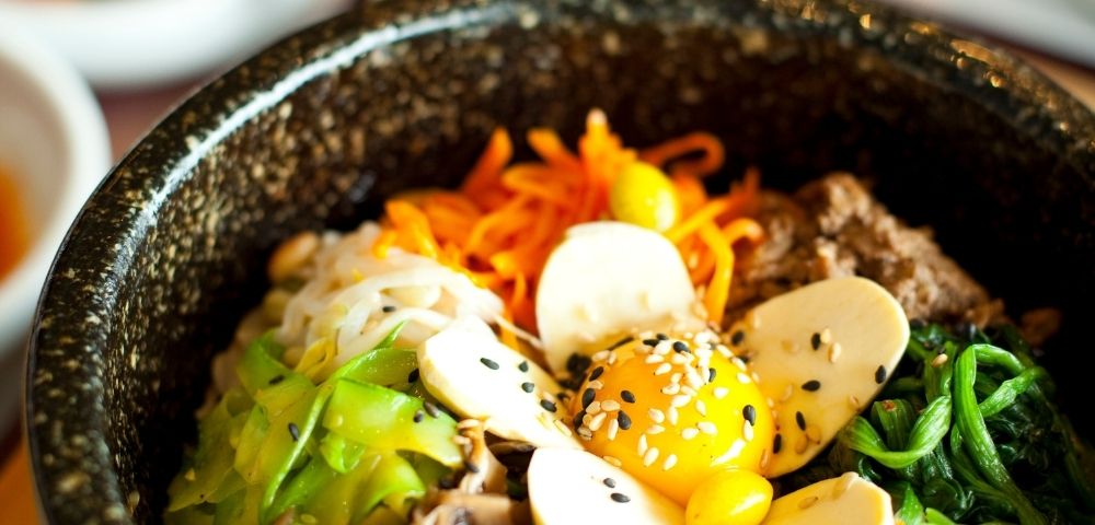 A close-up of a bibimbap bowl with assorted vegetables, sliced mushrooms, raw egg yolk, and sesame seeds on top.