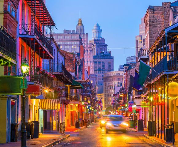 View of a bustling street lined with colorful buildings with wrought-iron balconies and gas lamps, illuminated by vibrant lights, under a twilight sky, with a car driving down the street.
