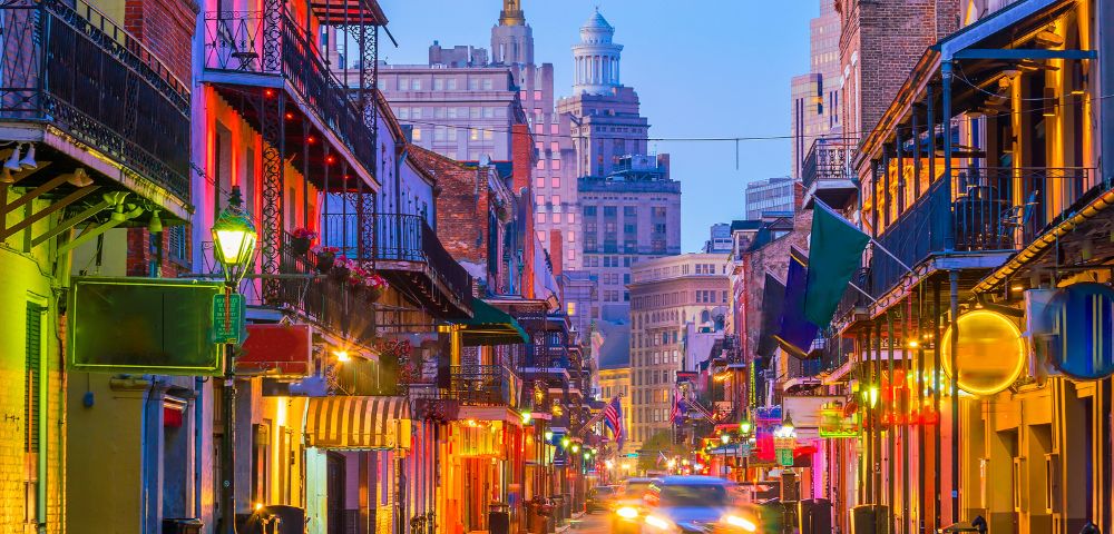 View of a bustling street lined with colorful buildings with wrought-iron balconies and gas lamps, illuminated by vibrant lights, under a twilight sky, with a car driving down the street.
